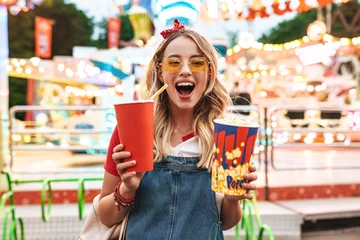 Keuken foto achterwand Amusementspark Afbeelding van een opgewonden charmante vrouw die popcorn en een kopje frisdrank vasthoudt tijdens het wandelen in het pretpark