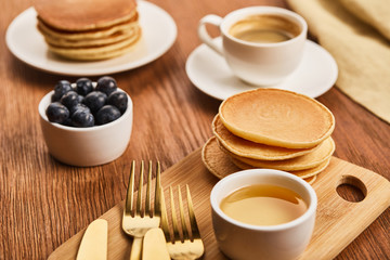 Fototapeta na wymiar selective focus of pancakes near bowl with honey and blueberries, golden cutlery and cup of coffee