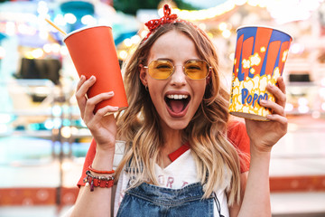 Afbeelding van een vrolijke charmante vrouw die popcorn en een kopje frisdrank vasthoudt tijdens het wandelen in het pretpark