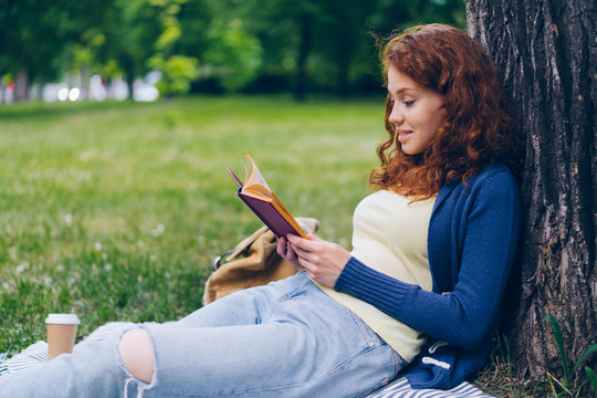 Good-looking young lady reading book and smiling sitting on lawn in park on beautiful summer day relaxing in recreational area. People, hobby and culture concept.