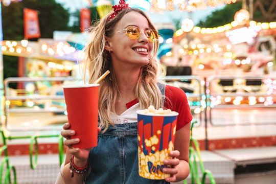 Image of blonde charming woman holding popcorn and soda paper cup while walking in amusement park