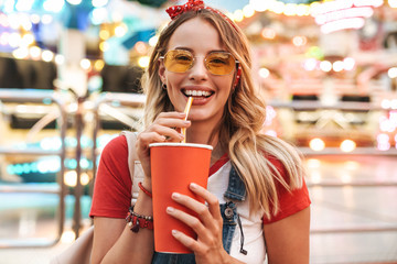 Image of young attractive woman drinking soda beverage from paper cup while walking in amusement...