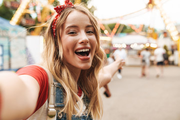 Image of joyful blonde woman rejoicing and taking selfie photo at amusement park