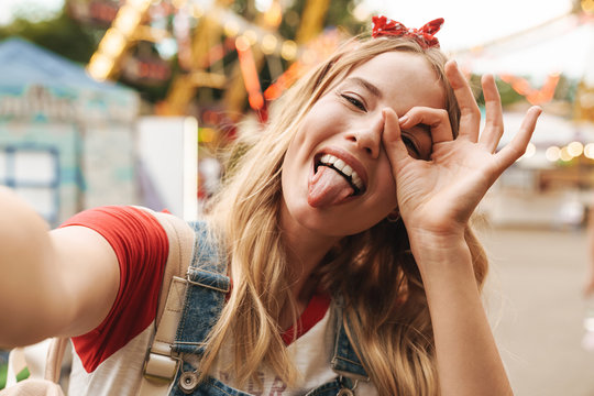 Image of happy blonde woman showing ok sign and taking selfie photo at amusement park