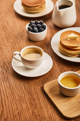 Fototapeta na wymiar selective focus of cup of coffee on saucer near plate with pancakes and bowls with honey and blueberries on wooden surface