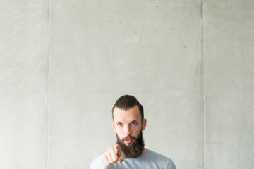 Judge and blame. Portrait of sceptic hipster guy standing over gray wall background, pointing finger at you. Copy space.