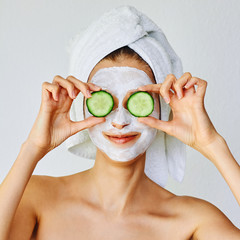 Beautiful young woman with facial mask on her face holding slices of cucumber. Skin care and...