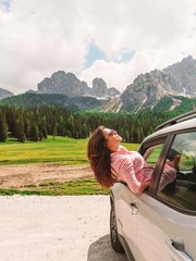 Brunette girl with long hair leans out of the car window against the background of the dolomite Mountains