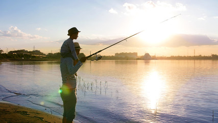 A man with a child on his shoulders fishing on the river bank at sunset