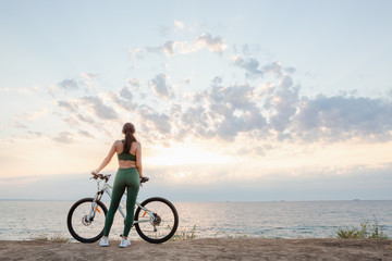 Fototapeta premium Beautiful young brunette woman resting after bicycle ride at sunrise. Model listening to music with wireless earbuds in the morning.