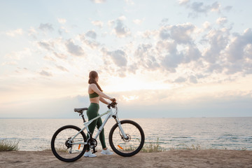 Fototapeta na wymiar Beautiful young brunette woman resting after bicycle ride at sunrise. Model listening to music with wireless earbuds in the morning.