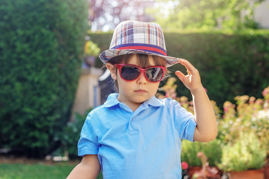 Portrait of funny little boy in sunglasses and plaid hat outdoors in garden at sunny summer day.  Summer lifestyle. Waving hello or goodbye