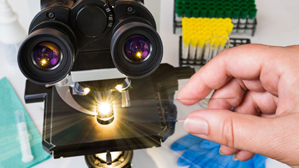 Laboratory optical microscope. Human hand holding a specimen on glass slide. Black medical scope, eyepieces, shining light rays, test tubes in rack, syringe and glove. Clinical analysis. Health care.
