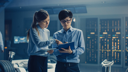 Male and Female Automobile Design Electric Car Engineers Having Conversation, Use Clipboard to...