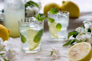 Bottle and glasses of cold lemon drink, ice, mint leaves, slices, white apple flowers on linen tablecloth. Close-up, copy space