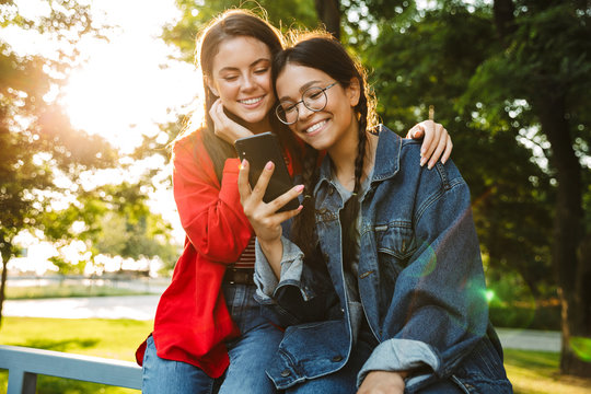 Image of two smiling girls using cellphone and hugging while sitting on railing in green park