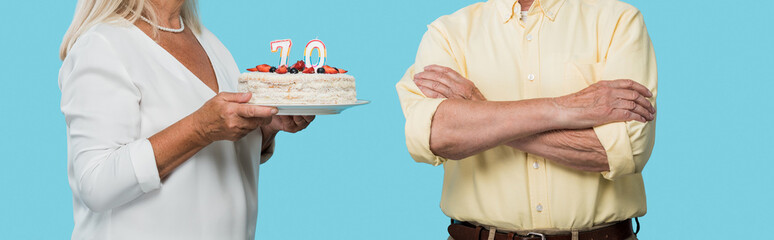 panoramic shot retired woman holding birthday cake near husband with crossed arms isolated on blue