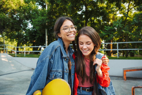 Image of two delighted girls smiling and hugging together while holding skateboard in skate park