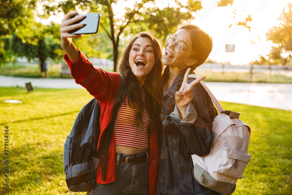 Sticker Image of two excited girls taking selfie photo on cellphone and gesturing peace sign while walking in green park