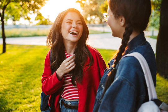 Image of two laughing girls students talking and looking at each other while walking in green park