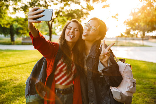 Image of two funny girls with tongues hanging out taking selfie photo on cellphone and gesturing peace sign while walking