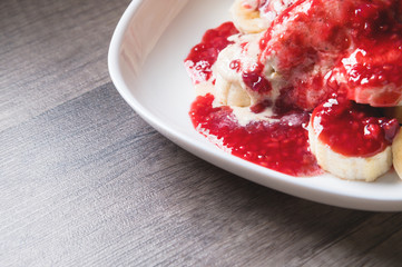 Closeup Vanilla ice cream poured with strawberry or raspberry jam with sliced banana slices on a white plate on a dark wooden table. You can do this at home on vacation
