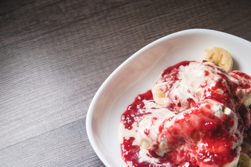 Closeup Vanilla ice cream poured with strawberry or raspberry jam with sliced banana slices on a white plate on a dark wooden table. You can do this at home on vacation