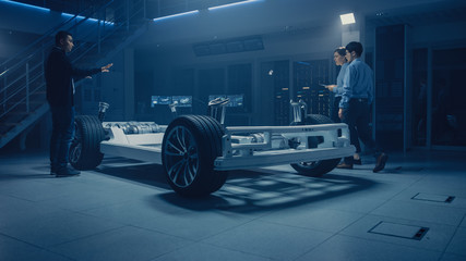 Automobile Design Engineers Talking while Working on Electric Car Chassis Prototype. In Automotive Innovation Facility Concept Vehicle Frame Includes Tires, Suspension, Engine and Battery