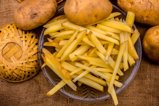 Raw cut french fries in a transparent glass plate along with raw potato with it on jute bag's surface.