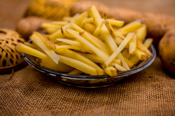 Raw cut french fries in a transparent glass plate along with raw potato with it on jute bag's surface.