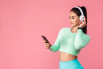 Pleased beautiful young sports fitness woman isolated over pink wall background using mobile phone listening music with headphones. - Powered by Adobe