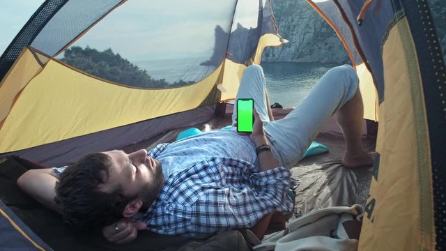 The solar panel attached to the tent. The man sitting next to mobile phone charges from the sun. A young guy lies in a camp tent, holding a smartphone with a green screen. Camp tent stands on beach