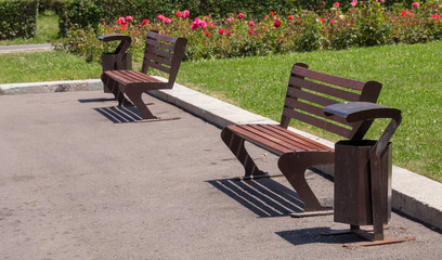 urn next to a street bench. Sidewalk with wooden bench and dumpster. Black metal wastebasket near the bench on the street