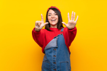 Young Mexican woman with overalls over yellow wall counting seven with fingers