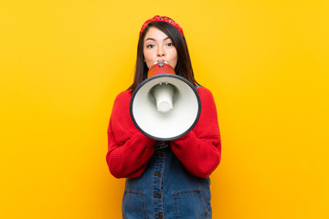 Young Mexican woman with overalls over yellow wall shouting through a megaphone