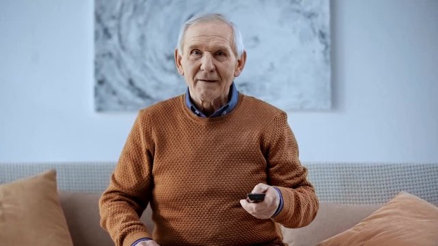 senior man sitting on sofa and holding remote controller while watching tv at home 