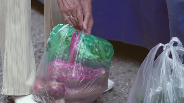 Elderly woman's hands tying various plastic carrying bags filled with vegetables together at a farmers street market in slow motion.