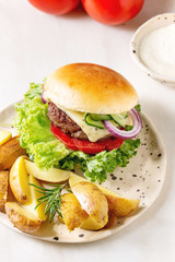 Homemade fast food burger classic hamburger or cheeseburger with beef, salad, cheese and tomato served on ceramic plate with baked country potatoes and sauce on white marble kitchen table.