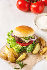 Homemade fast food burger classic hamburger or cheeseburger with beef, salad, cheese and tomato served on paper with baked country potatoes and sauce on white marble kitchen table.