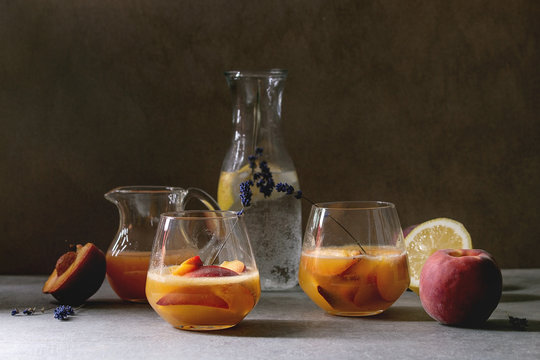 Peach cold lemonade or Bellini cocktail with lavender flowers served in two glasses with pitcher of pesh juice and lemon soda water on dark texture table. Homemade summer drink.