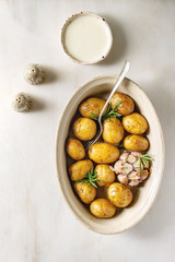 Young baked potatoes with garlic and rosemary in ceramic baking dish, served with cheese sauce, salt and pepper over white marble background. Flat lay, space