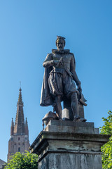 Fototapeta na wymiar Bruges, Flanders, Belgium - June 17, 2019: Simon Stevin statue with spire of OLV Cathedral in back under blue sky. Some green foliage. Pigeon on his head.