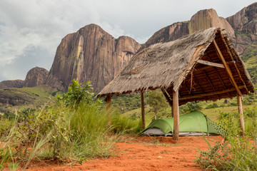 Small hiking tent pitched under an open thatched hut in the sacred forest, Tsaranoro valley, Madagascar. Famous rock formation mountain ridge on background 