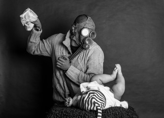 Young Dad cleaning his baby's dirty ass, changing the stinky diaper in a gas mask, fatherhood and humor..Father changes the baby's diapers on a changing table, on a dark, single track background. - obrazy, fototapety, plakaty