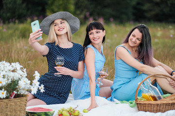 Three young women, in blue dresses and hats sit on a plaid and take pictures on a smartphone. Outdoor picnic on the grass in the forest. Delicious food in a picnic basket and wine.