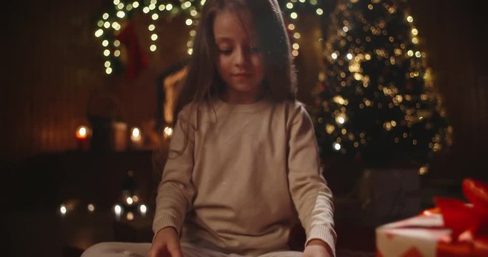 Little caucasian girl sitting near christmas tree in decorated room and opening her gift with something special - holidays and celebrations, christmas spirit concept close up 4k footage