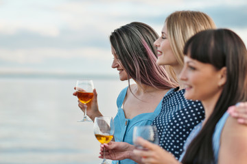 Three young attractive women, in blue dresses are walking along the seashore. Girlfriends communicate, laugh and drink wine.