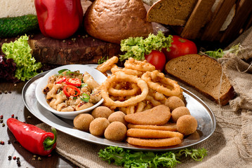 Assorted beer snacks: onion rings, cheese balls, chips etc on a iron tray on a dark wooden background among vegetables.