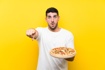 Young handsome man holding a pizza over isolated yellow wall surprised and pointing front