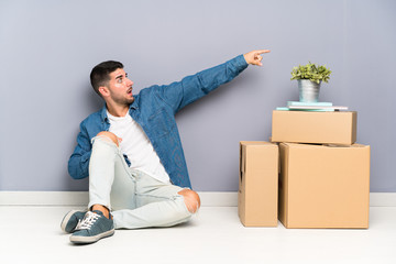 Handsome young man moving in new home among boxes pointing finger to the side